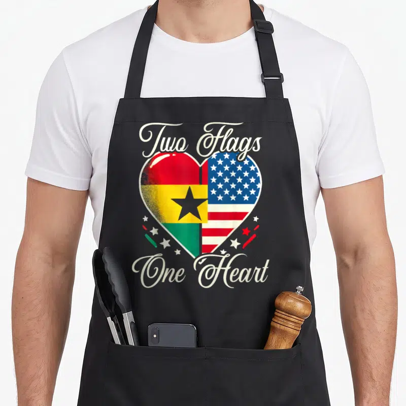 Ghanaian ghana american flag apron - Heart split into Ghana and USA flags with Two Flags One Heart script — shown on a black cotton canvas apron against a white background