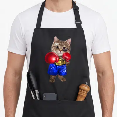 Boxing cat apron - Realistic brown tabby cat wearing red boxing gloves, blue shorts, and a gold CHAMPION belt — shown on a black cotton canvas apron against a white background