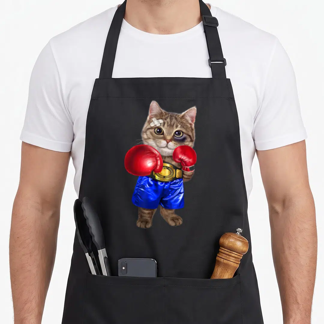 Boxing cat apron - Realistic brown tabby cat wearing red boxing gloves, blue shorts, and a gold CHAMPION belt — shown on a black cotton canvas apron against a white background