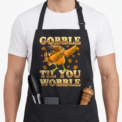 Til thanksgiving apron - Cartoon turkey dabbing with top hat and sunglasses surrounded by leaves and split text — shown on a black cotton canvas apron against a white background