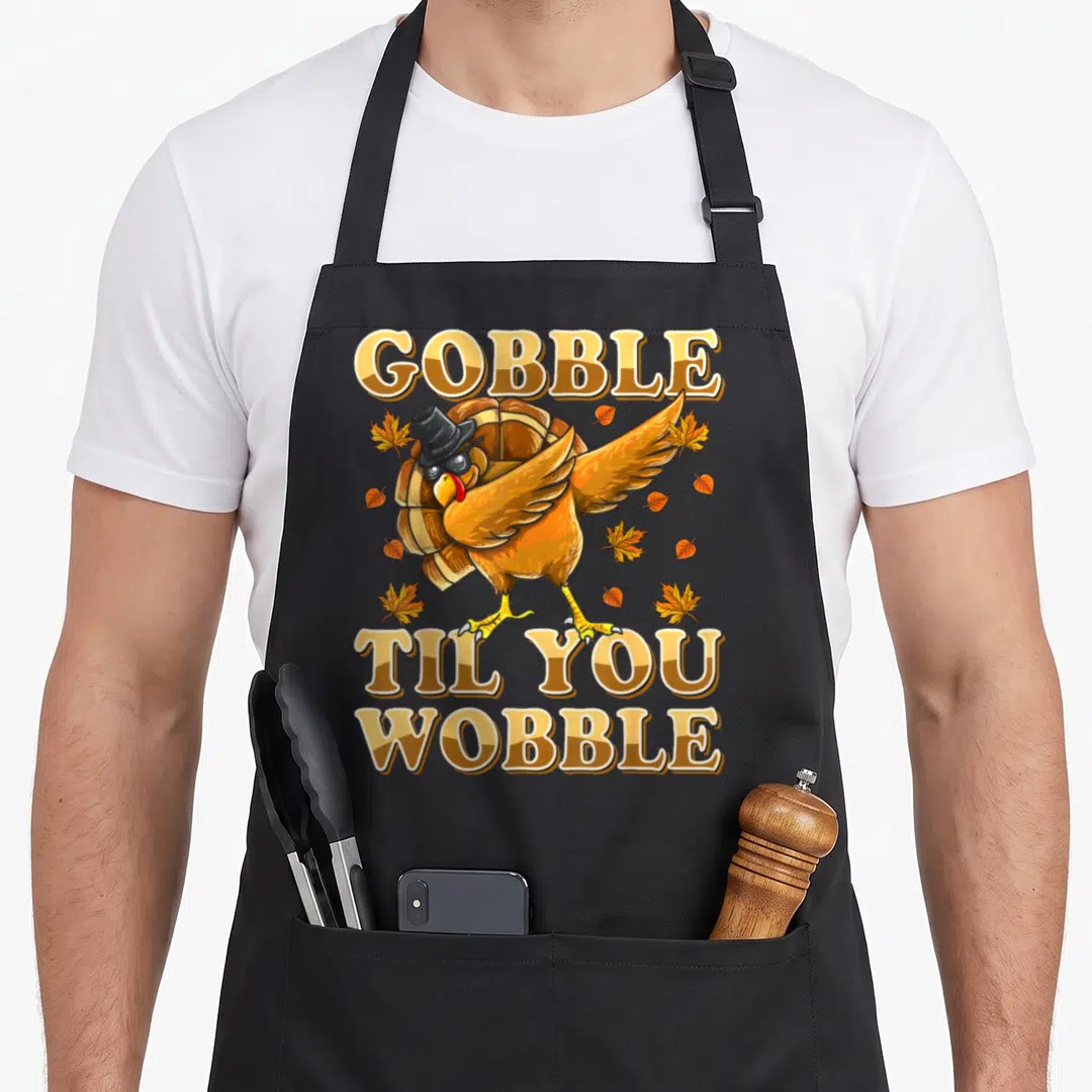 Til thanksgiving apron - Cartoon turkey dabbing with top hat and sunglasses surrounded by leaves and split text — shown on a black cotton canvas apron against a white background
