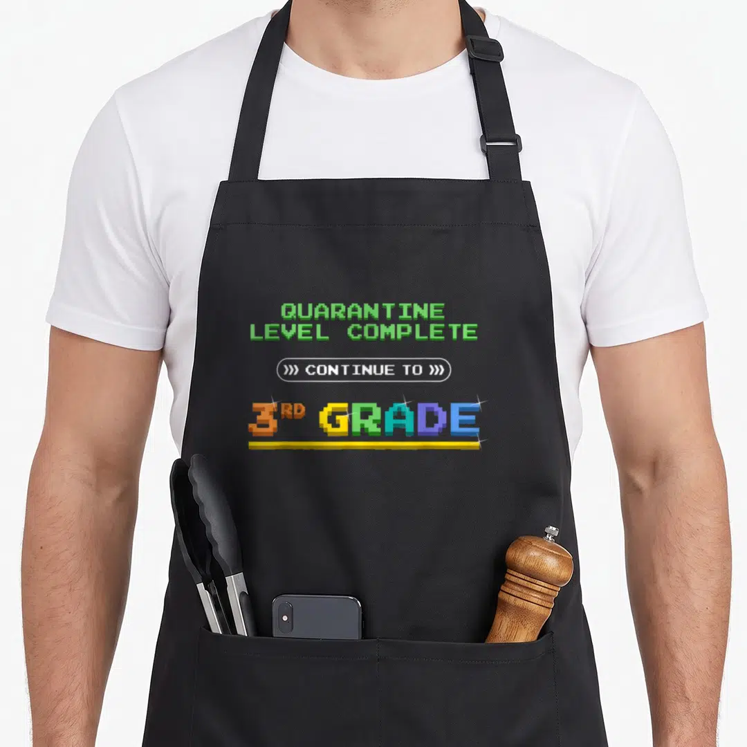Complete quarantine aprons - Pixel art text QUARANTINE LEVEL COMPLETE CONTINUE TO 3RD GRADE in green orange yellow blue — shown on a black cotton canvas apron against a white background