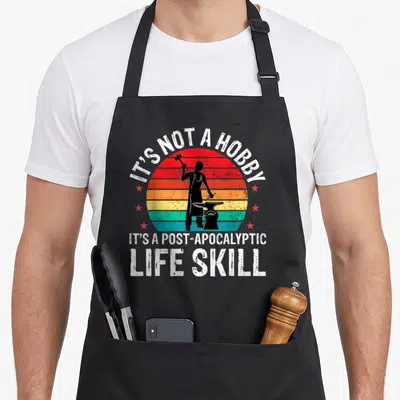 Anvil blacksmith apron - Blacksmith silhouette on striped sun with IT'S NOT A HOBBY text — shown on a black cotton canvas apron against a white background