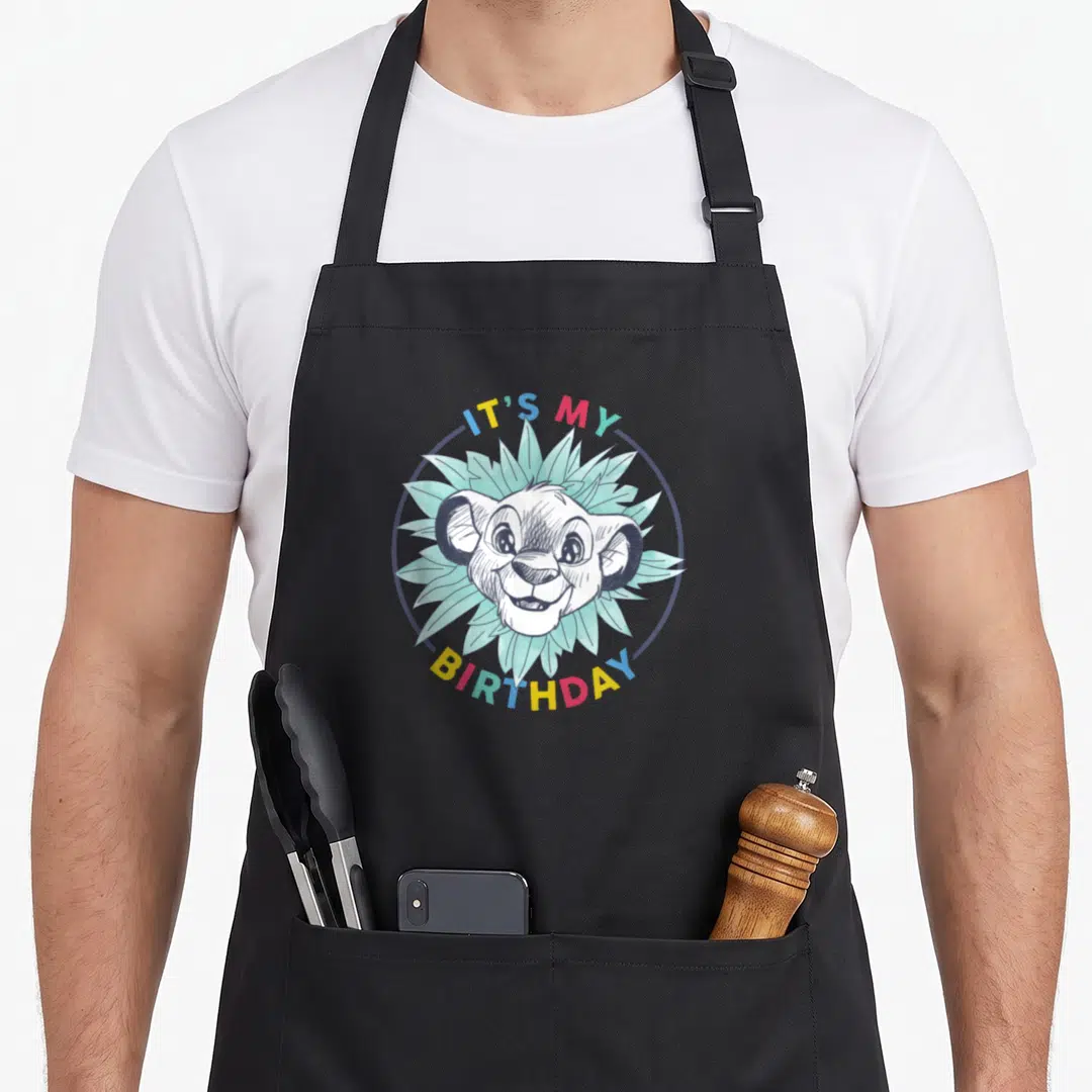 Simba floral aprons - Cartoon lion cub head surrounded by leaves and IT'S MY BIRTHDAY text in rainbow colors — shown on a black cotton canvas apron against a white background