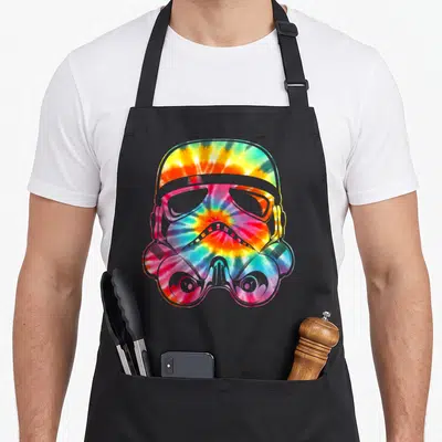 Face tie apron - Tie-dye stormtrooper helmet in red yellow green and blue on black fabric — shown on a black cotton canvas apron against a white background