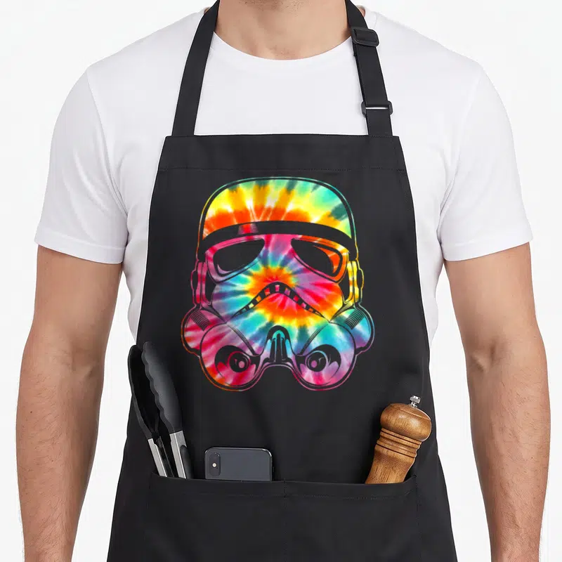 Face tie apron - Tie-dye stormtrooper helmet in red yellow green and blue on black fabric — shown on a black cotton canvas apron against a white background