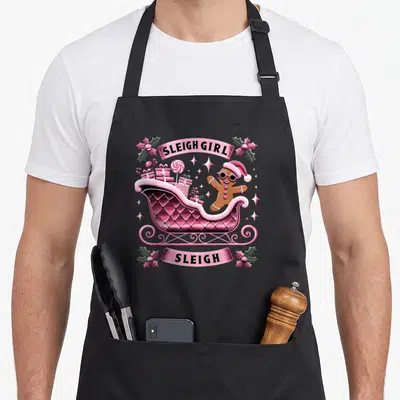 Groovy gingerbread apron - Pink sleigh with gingerbread man and SLEIGH GIRL text banners — shown on a black cotton canvas apron against a white background