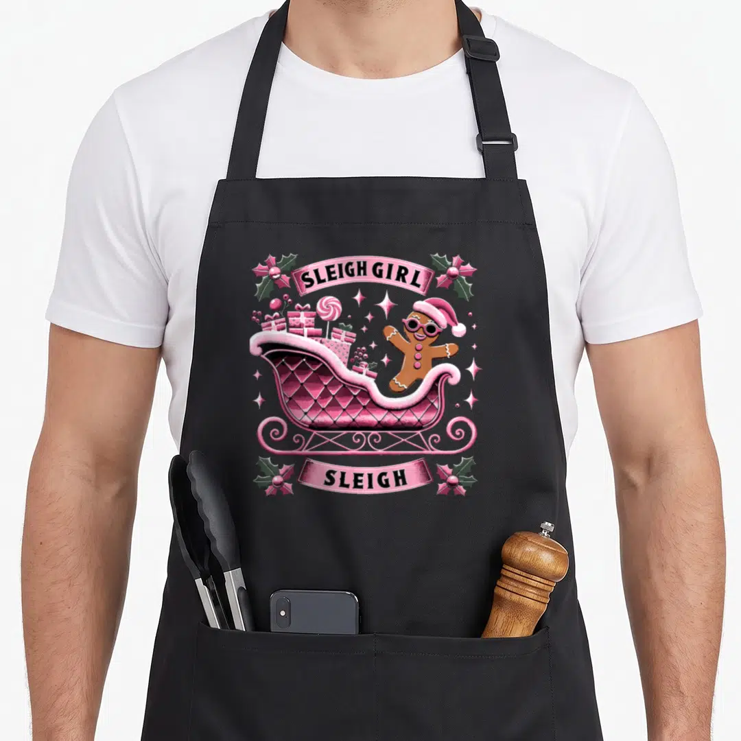 Groovy gingerbread apron - Pink sleigh with gingerbread man and SLEIGH GIRL text banners — shown on a black cotton canvas apron against a white background