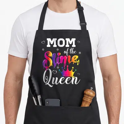 Slime crown queen of baking apron - MOM of the Slime Queen text with dripping rainbow letters, crown, stars and hearts — shown on a black cotton canvas apron against a white background
