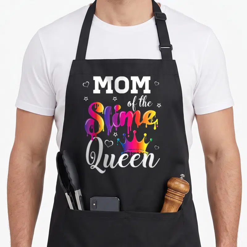 Slime crown queen of baking apron - MOM of the Slime Queen text with dripping rainbow letters, crown, stars and hearts — shown on a black cotton canvas apron against a white background