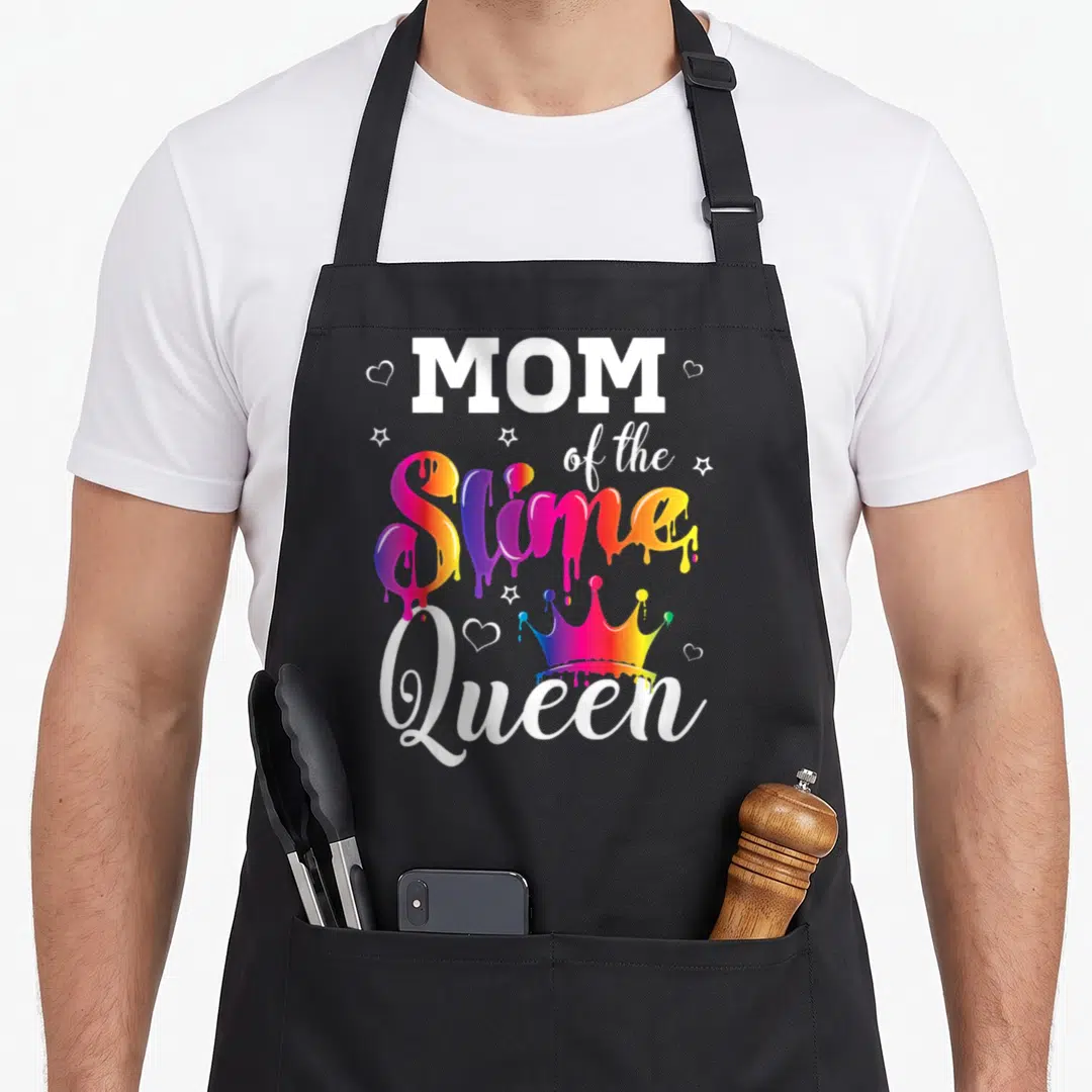 Slime crown queen of baking apron - MOM of the Slime Queen text with dripping rainbow letters, crown, stars and hearts — shown on a black cotton canvas apron against a white background