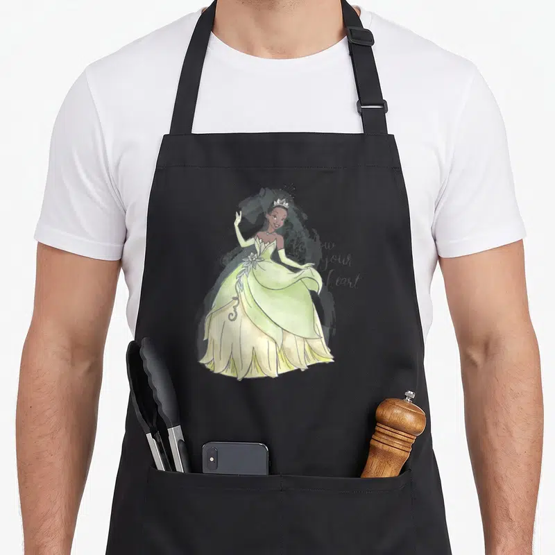 Follow princess apron - Watercolor princess in green gown with follow your heart script text — shown on a black cotton canvas apron against a white background
