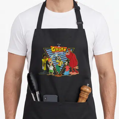 Movie crew apron - A Goofy Movie title text above five cartoon characters in front of a yellow and blue striped triangle — shown on a black cotton canvas apron against a white background