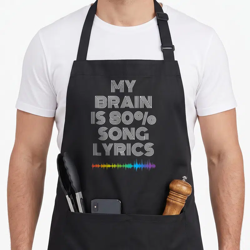Brain singing addict apron - White striped text MY BRAIN IS 80% SONG LYRICS above multicolor audio wave — shown on a black cotton canvas apron against a white background