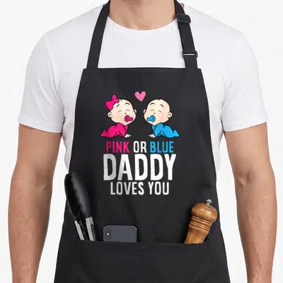 Gender reveal daddy apron - Two cartoon babies in pink and blue outfits with PINK OR BLUE DADDY LOVES YOU text — shown on a black cotton canvas apron against a white background