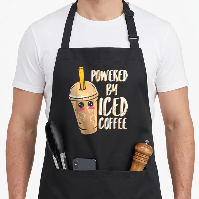 Iced anime apron - Cartoon iced coffee cup with yellow straw next to stacked POWERED BY ICED COFFEE text in light brown — shown on a black cotton canvas apron against a white background