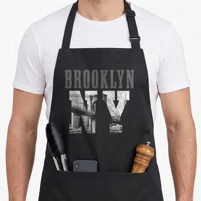 Vintage nyc new york apron - BROOKLYN NY text with Brooklyn Bridge photo filling the letters in distressed grey — shown on a black cotton canvas apron against a white background