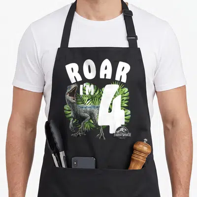 Jurassic birthday apron - Velociraptor and ROAR I'M 4 text over green palm leaves — shown on a black cotton canvas apron against a white background