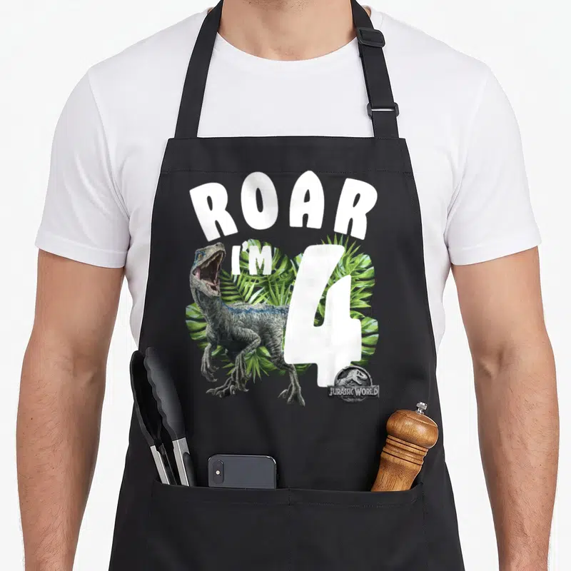 Jurassic birthday apron - Velociraptor and ROAR I'M 4 text over green palm leaves — shown on a black cotton canvas apron against a white background