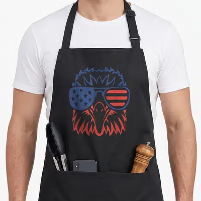 Eagle 4th of july apron - Cartoon eagle head with blue and red sunglasses featuring stars and stripes pattern — shown on a black cotton canvas apron against a white background