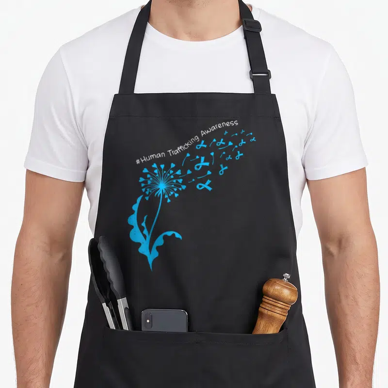 Dandelion human trafficking awareness apron - Blue dandelion seeds turn into hearts and ribbons under white arched text — shown on a black cotton canvas apron against a white background
