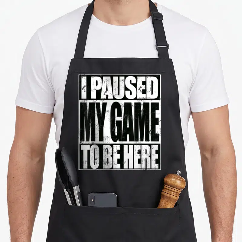 Paused sarcastic apron - Stacked white and black text reading I Paused My Game To Be Here with distressed finish — shown on a black cotton canvas apron against a white background