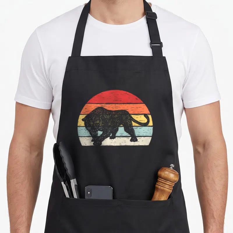 Funny catch some rays apron - Retro black panther silhouette over red orange yellow blue cream and grey horizontal sun — shown on a black cotton canvas apron against a white background