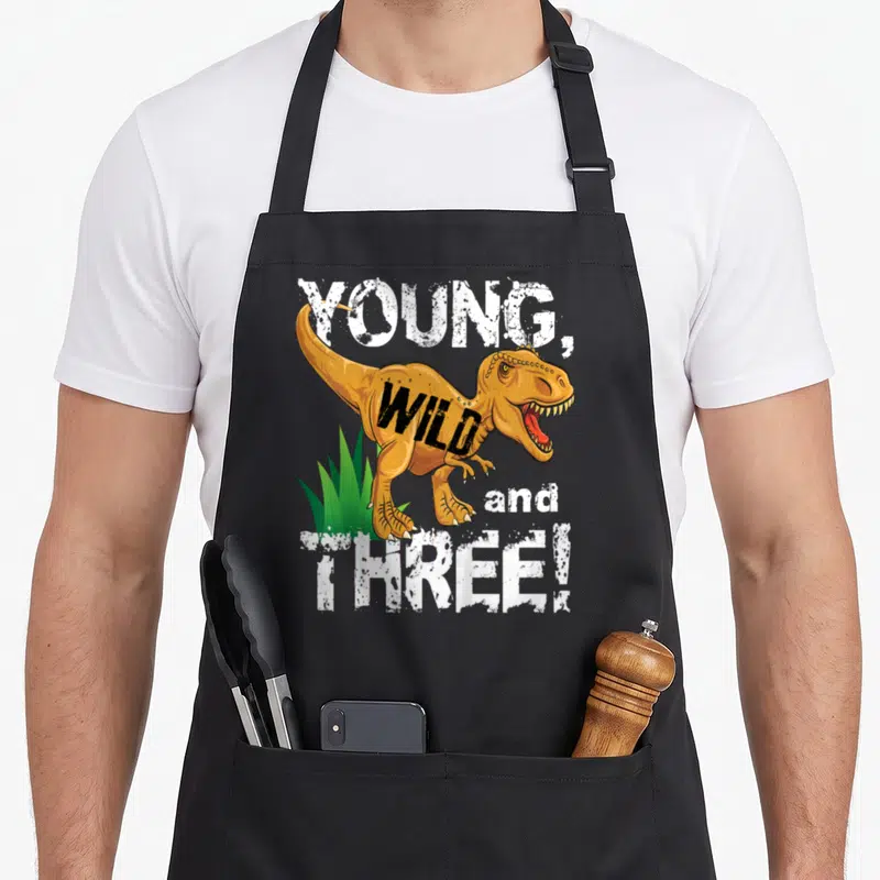 Young dinosaur apron - Cartoon T-Rex with YOUNG WILD and THREE text in orange white and green — shown on a black cotton canvas apron against a white background