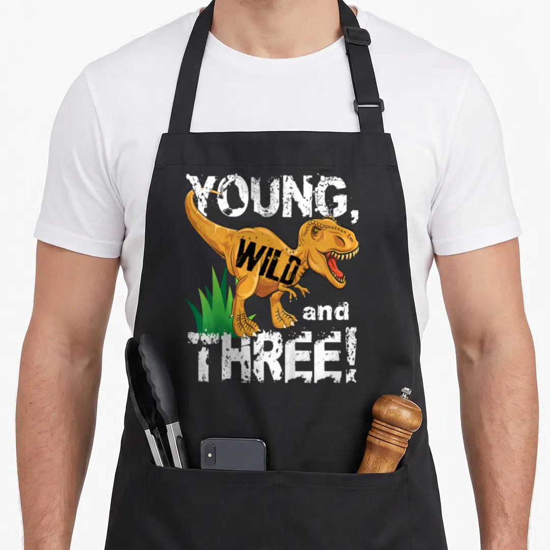 Young dinosaur apron - Cartoon T-Rex with YOUNG WILD and THREE text in orange white and green — shown on a black cotton canvas apron against a white background