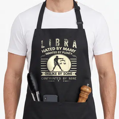 Hated libra apron - Distressed white text reading Hated By Many above a man holding scales in a circle — shown on a black cotton canvas apron against a white background