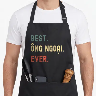 Vietnamese grandpa apron - Stacked text reading BEST ONG NGOAI EVER in green yellow and red distressed font — shown on a black cotton canvas apron against a white background