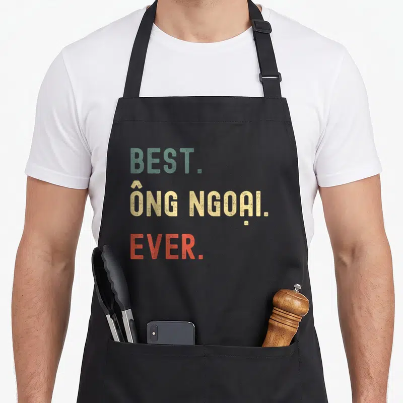 Vietnamese grandpa apron - Stacked text reading BEST ONG NGOAI EVER in green yellow and red distressed font — shown on a black cotton canvas apron against a white background