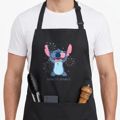Sparkle 4th of july apron - Stitch wearing US flag sunglasses surrounded by fireworks with BORN TO SPARKLE text below — shown on a black cotton canvas apron against a white background