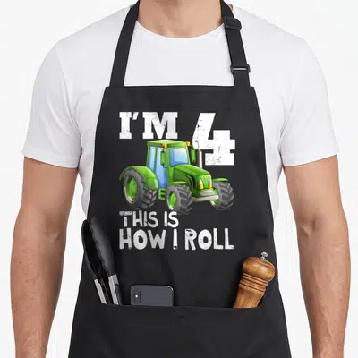 4th farm apron - Green cartoon tractor with white text reading I'M 4 THIS IS HOW I ROLL — shown on a black cotton canvas apron against a white background