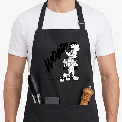 Cute paint apron - Woody Woodpecker holding two brushes next to dripping graffiti text WOODY — shown on a black cotton canvas apron against a white background