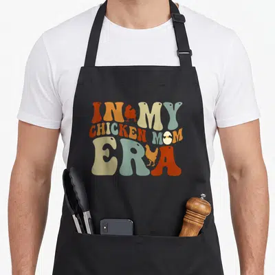 Era chicken apron - Stacked text IN MY CHICKEN MOM ERA with chicken silhouettes and chick in egg, orange tan blue grey — shown on a black cotton canvas apron against a white background