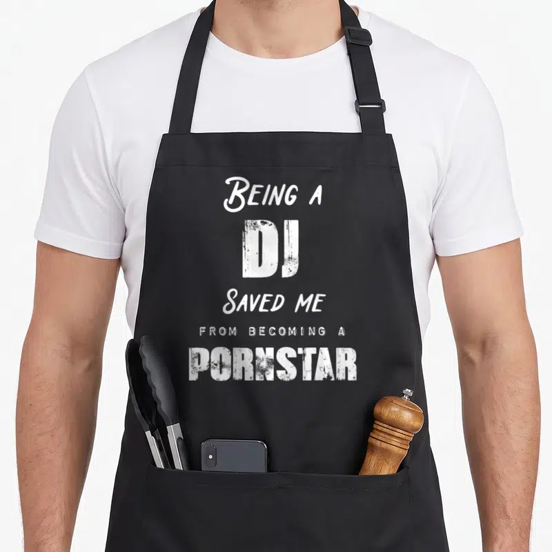 Saved nurse apron - White stacked text reading Being a DJ Saved Me from Becoming a Pornstar — shown on a black cotton canvas apron against a white background