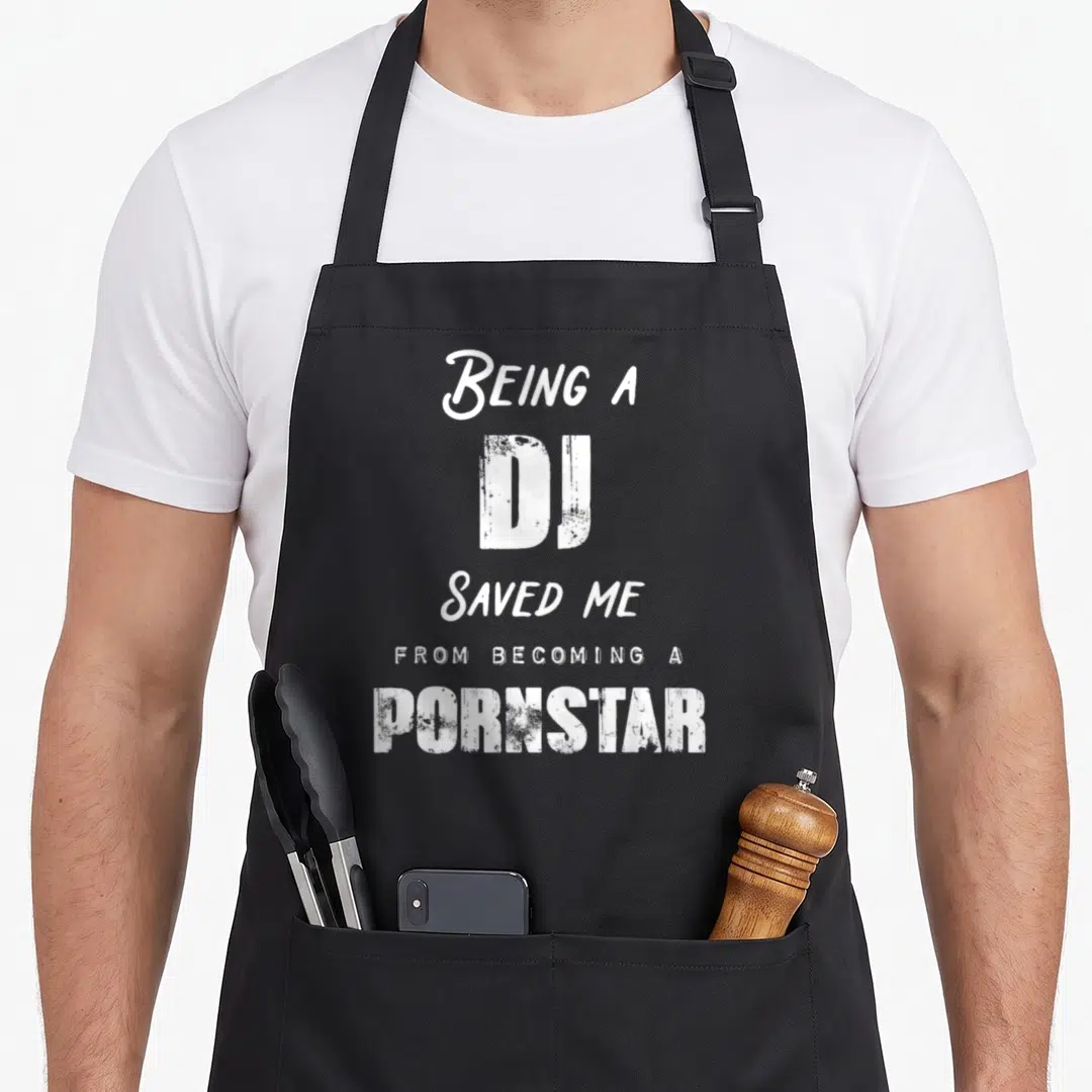 Saved nurse apron - White stacked text reading Being a DJ Saved Me from Becoming a Pornstar — shown on a black cotton canvas apron against a white background