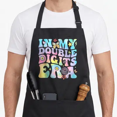 Double digit era birthday apron - Retro bubble text reading In My Double Digits Era with butterflies, disco ball — shown on a black cotton canvas apron against a white background