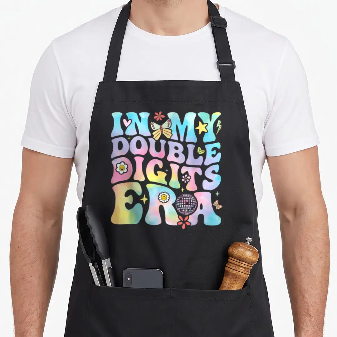Double digit era birthday apron - Retro bubble text reading In My Double Digits Era with butterflies, disco ball — shown on a black cotton canvas apron against a white background