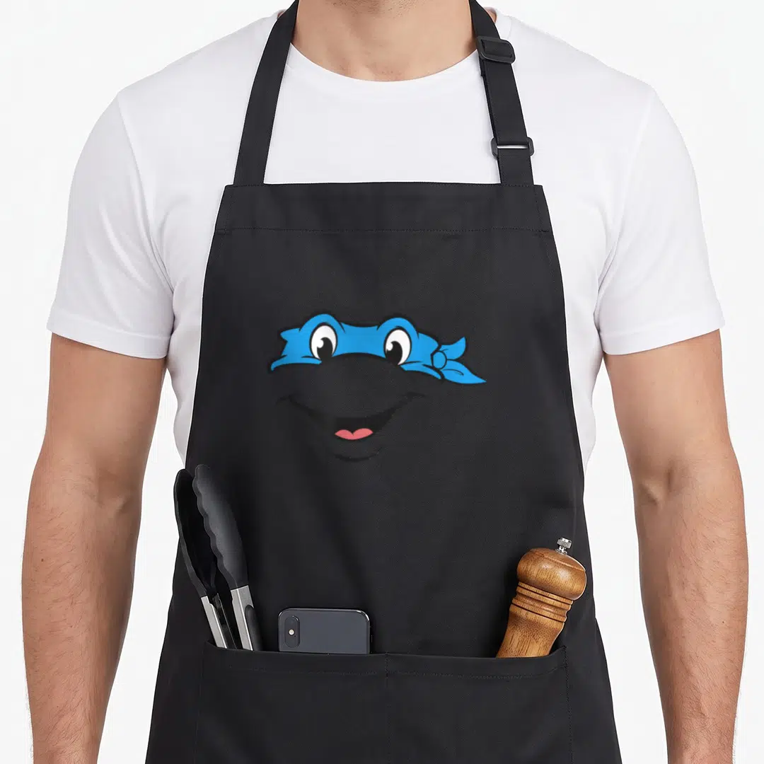 Leonardo face apron - Cartoon grey turtle face wearing a blue bandana mask with a pink tongue visible in a wide smile — shown on a black cotton canvas apron against a white background