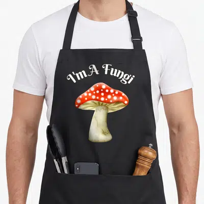Humorous mushroom apron - I'm A Fungi text over red spotted mushroom illustration — shown on a black cotton canvas apron against a white background
