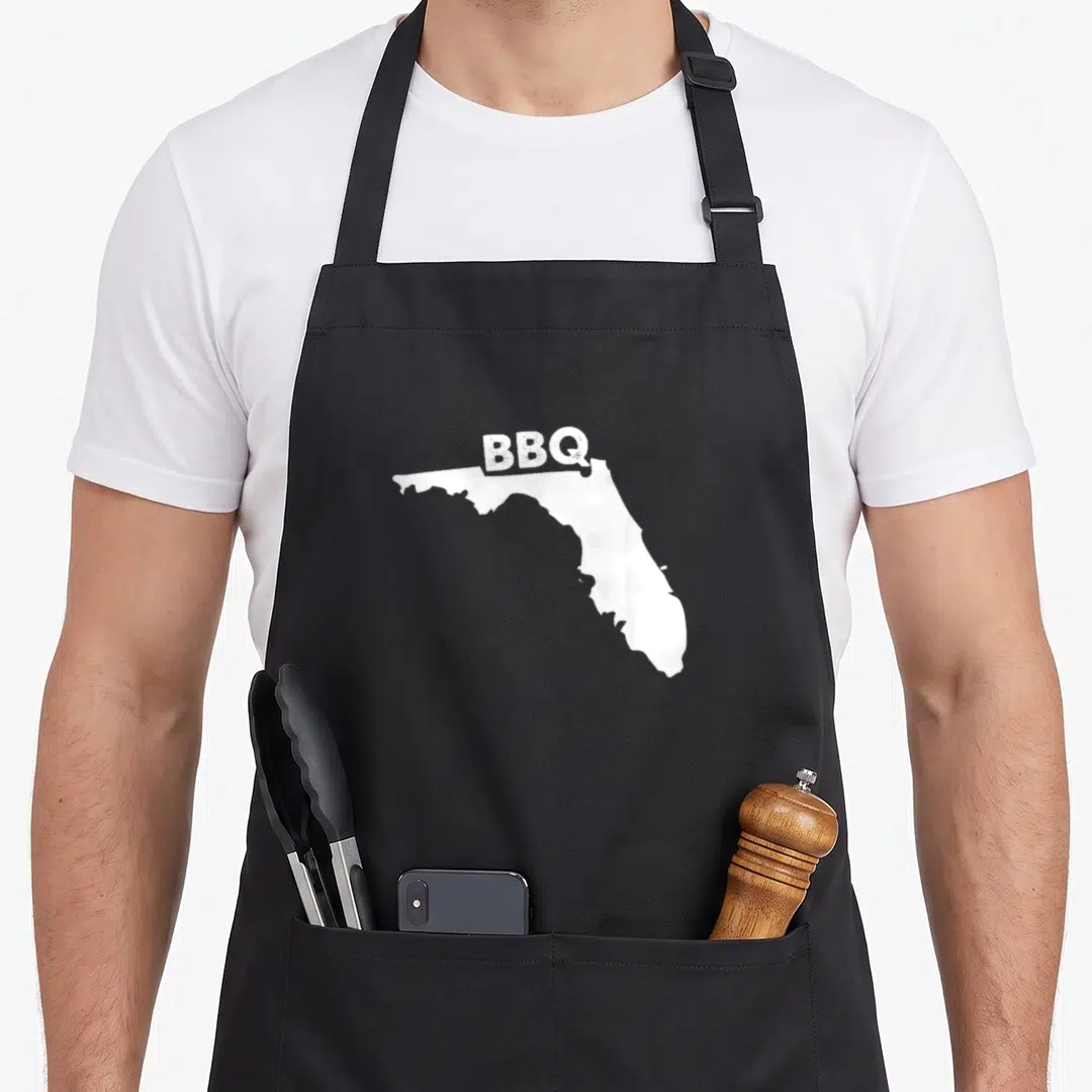 Florida barbecue apron - White Florida state silhouette with BBQ text printed above the outline — shown on a black cotton canvas apron against a white background