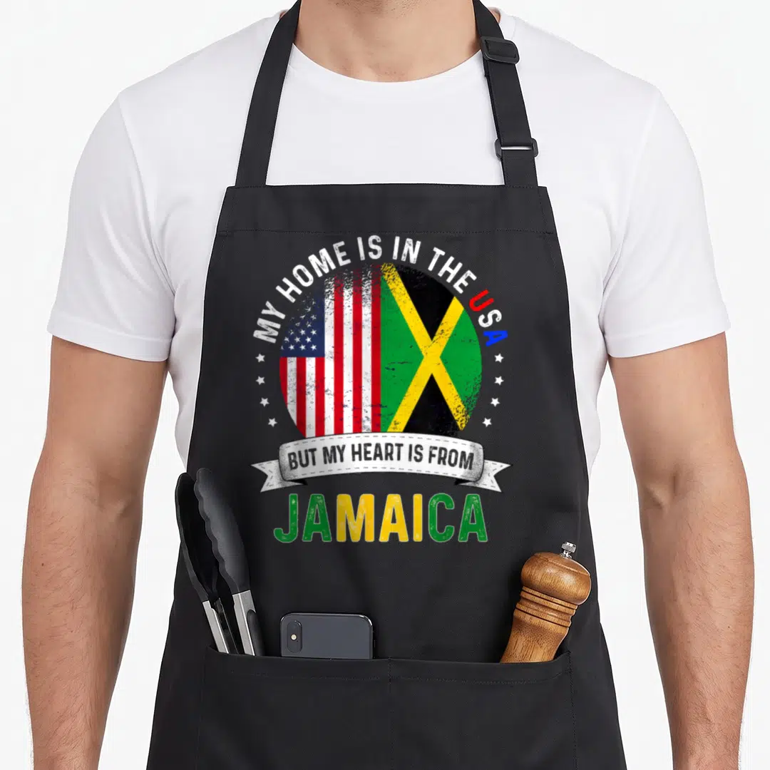 Jamaica patriots apron - Distressed oval with US and Jamaican flags and My Home Is In The USA But My Heart Is — shown on a black cotton canvas apron against a white background