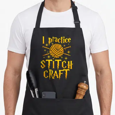 Practice craft apron - Yellow yarn ball with crossed needles surrounded by stars and lightning bolts between stacked — shown on a black cotton canvas apron against a white background