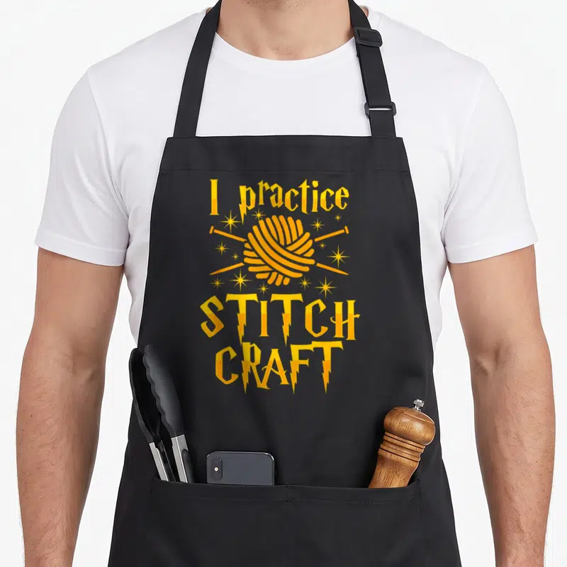 Practice craft apron - Yellow yarn ball with crossed needles surrounded by stars and lightning bolts between stacked — shown on a black cotton canvas apron against a white background
