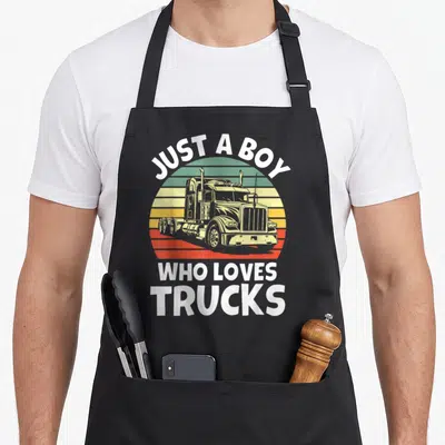 Trucker toddler semi truck apron - Retro sunset graphic with semi-truck and white text JUST A BOY WHO LOVES TRUCKS — shown on a black cotton canvas apron against a white background