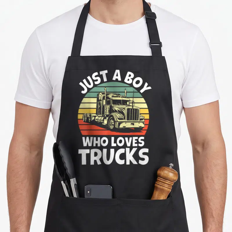 Trucker toddler semi truck apron - Retro sunset graphic with semi-truck and white text JUST A BOY WHO LOVES TRUCKS — shown on a black cotton canvas apron against a white background