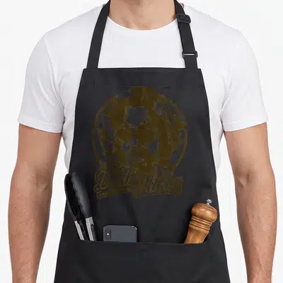 Negative dude apron - Distressed brown circle portrait of The Dude with script text The Dude Abides at bottom — shown on a black cotton canvas apron against a white background