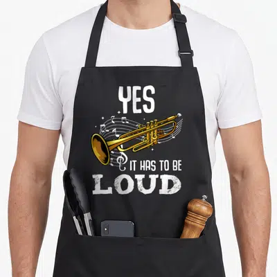 Das tablet apron - Gold trumpet with white musical notes and text YES IT HAS TO BE LOUD — shown on a black cotton canvas apron against a white background